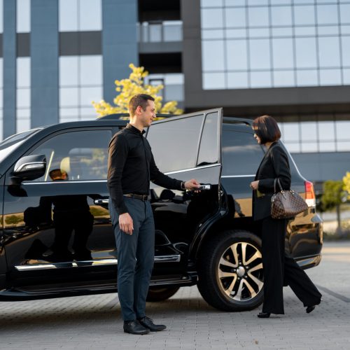 Chauffeur helps business couple to get in the car, opening a door of a luxury black taxi. Man and woman having a business trip together