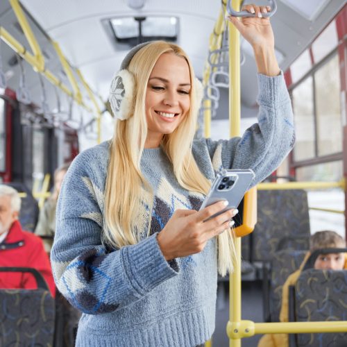 Front view of pritty blonde girl using phone smiling standing inside moving bus. Crop of female holding handhold with blur old bearded man and boy behind in urban transport. Concept of city life.
