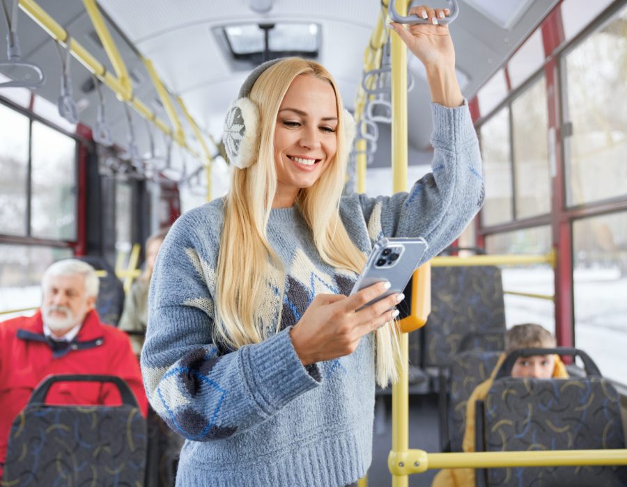 Front view of pritty blonde girl using phone smiling standing inside moving bus. Crop of female holding handhold with blur old bearded man and boy behind in urban transport. Concept of city life.
